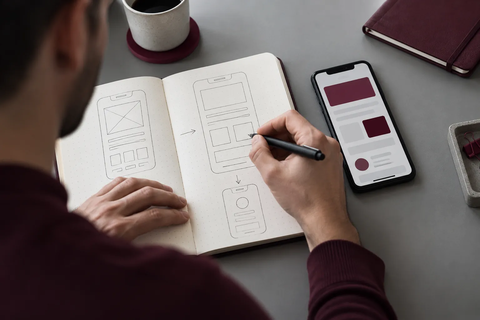 Mãos desenhando telas de um aplicativo em um caderno ao lado de um celular.